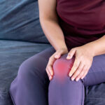 Older woman sitting on couch at home while experiencing knee pain caused by an osteoarthritis flare up