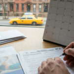 Close-up of hands filling out a New York State NF-2 No-Fault Benefits application form on a wooden desk. Beside the form is a physical therapy brochure and a desk calendar with "DEC 30" circled and labeled "- DEADLINE". Through a window in the background, a yellow taxi drives down a city street lined with brownstones.