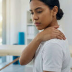 A woman experiencing shoulder and upper back pain in a physical therapy clinic setting.